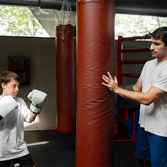 Child practicing punches on a heavy bag while a coach supervises and stabilizes the bag inside a boxing gym.
