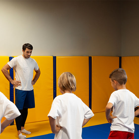 Coach instructing a group of children during a kids’ martial arts or fitness class on padded mats in a gym.