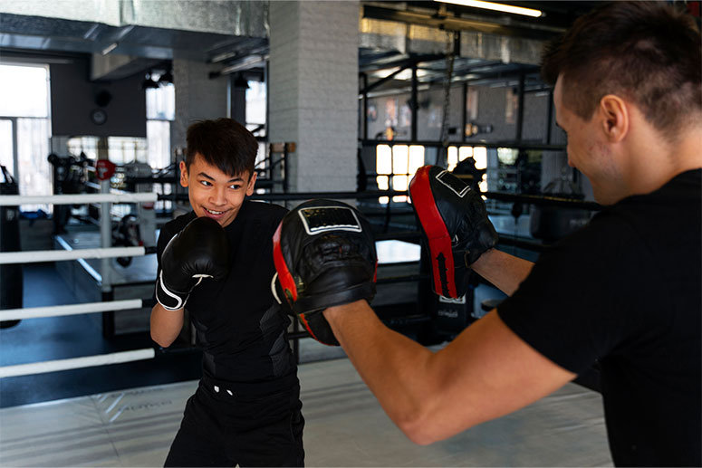 Young boxer practicing punching techniques with a trainer using focus mitts inside a boxing gym, wearing gloves and protective gear.