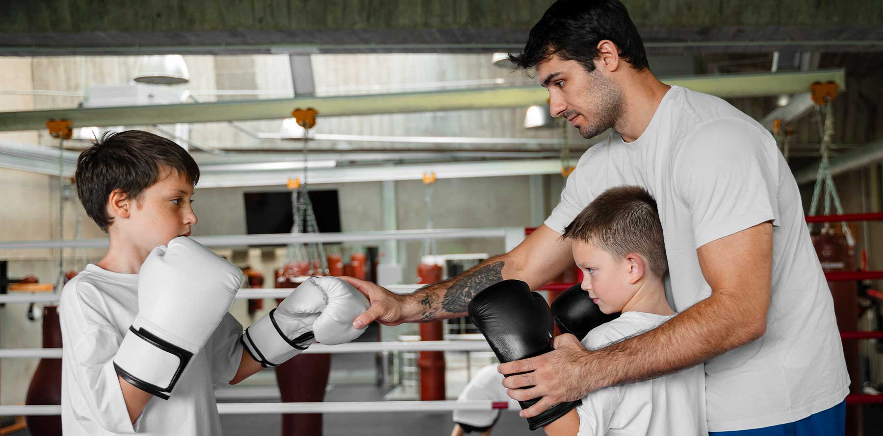 Coach guiding two children during boxing practice, correcting glove position and punching technique inside a boxing ring.